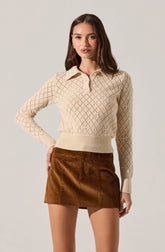 A woman is wearing a cream-colored knit sweater with a diamond pattern and a collar. The sweater has a zip-up neckline and ribbed cuffs and hem. She is also wearing a high-waisted mini skirt made of brown corduroy fabric.